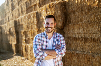 Handsome120 tys. zł na rozwój małych dospodarstw caucasian smiling farmer standing outdoors with arms crossed and looking at camera. In background are of hay.