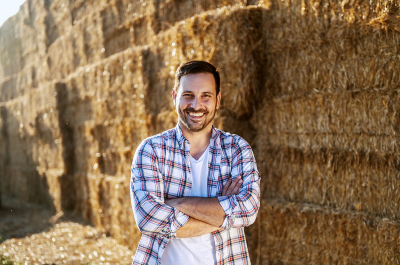 Handsome120 tys. zł na rozwój małych dospodarstw caucasian smiling farmer standing outdoors with arms crossed and looking at camera. In background are of hay.